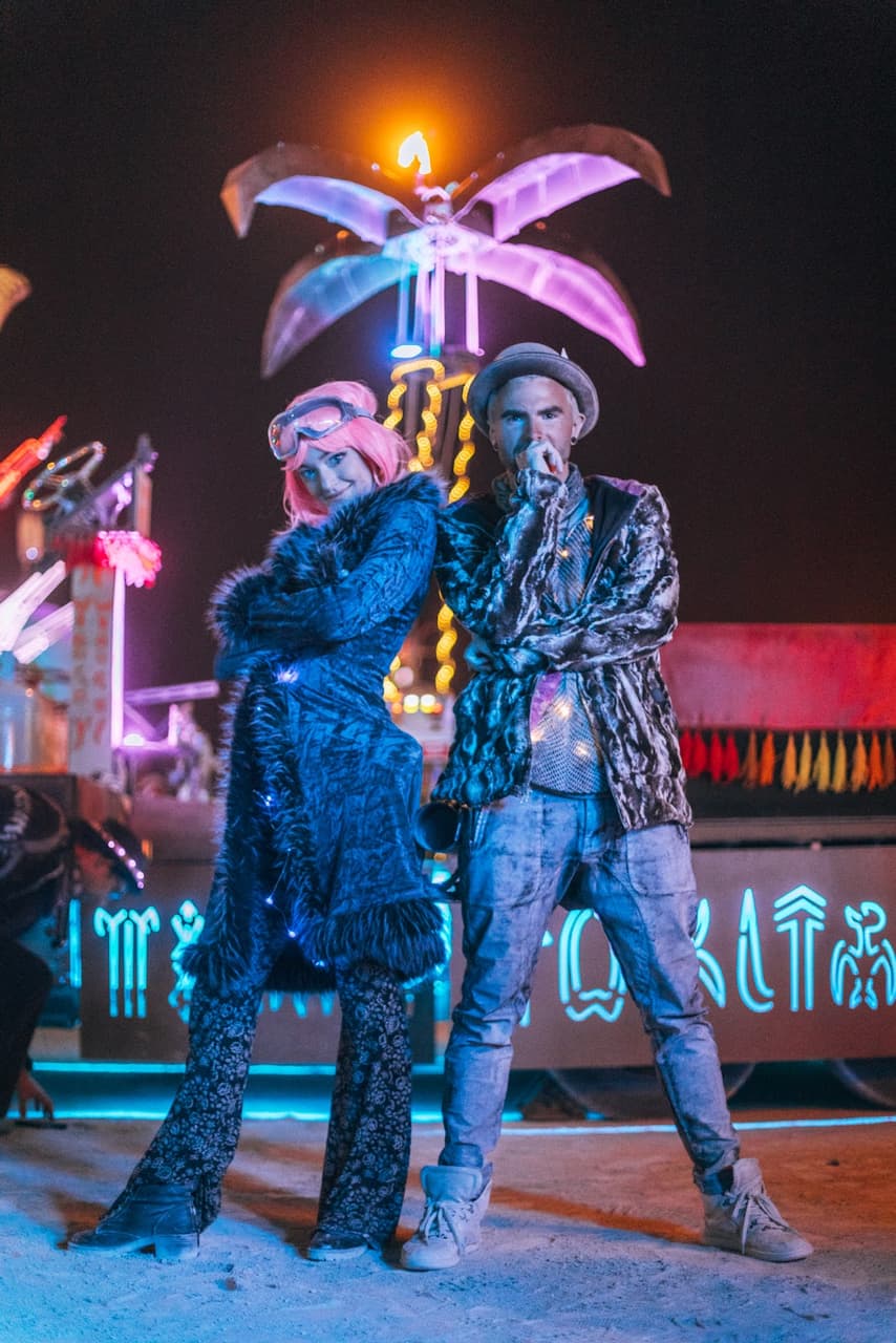 Two festively dressed individuals pose back-to-back at night amidst glowing art installations.