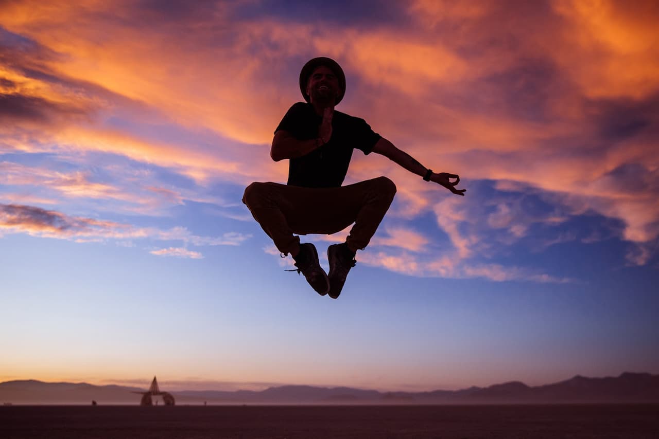 A silhouetted person joyfully leaps against a vibrant sunset sky over a vast, flat landscape.