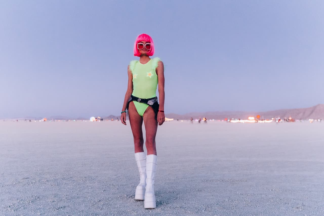 A person in a vibrant neon green bodysuit, pink wig, and heart-shaped sunglasses stands on a vast desert playa at twilight.