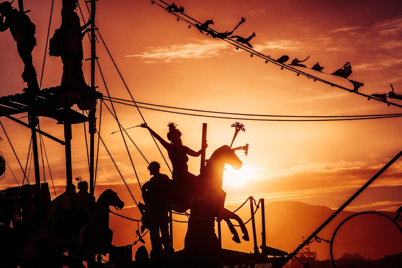 Silhouetted figures on an elaborate art installation stand against a vibrant orange and yellow sunset sky.