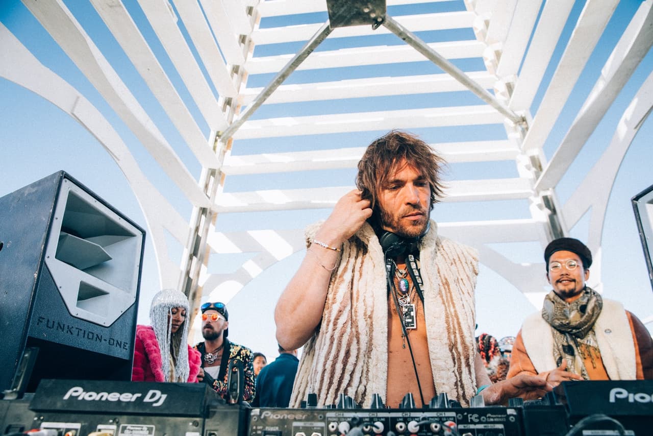 A focused DJ with shaggy hair and a fur-trimmed vest adjusts his headphones while mixing music at an outdoor event under a striking white ge