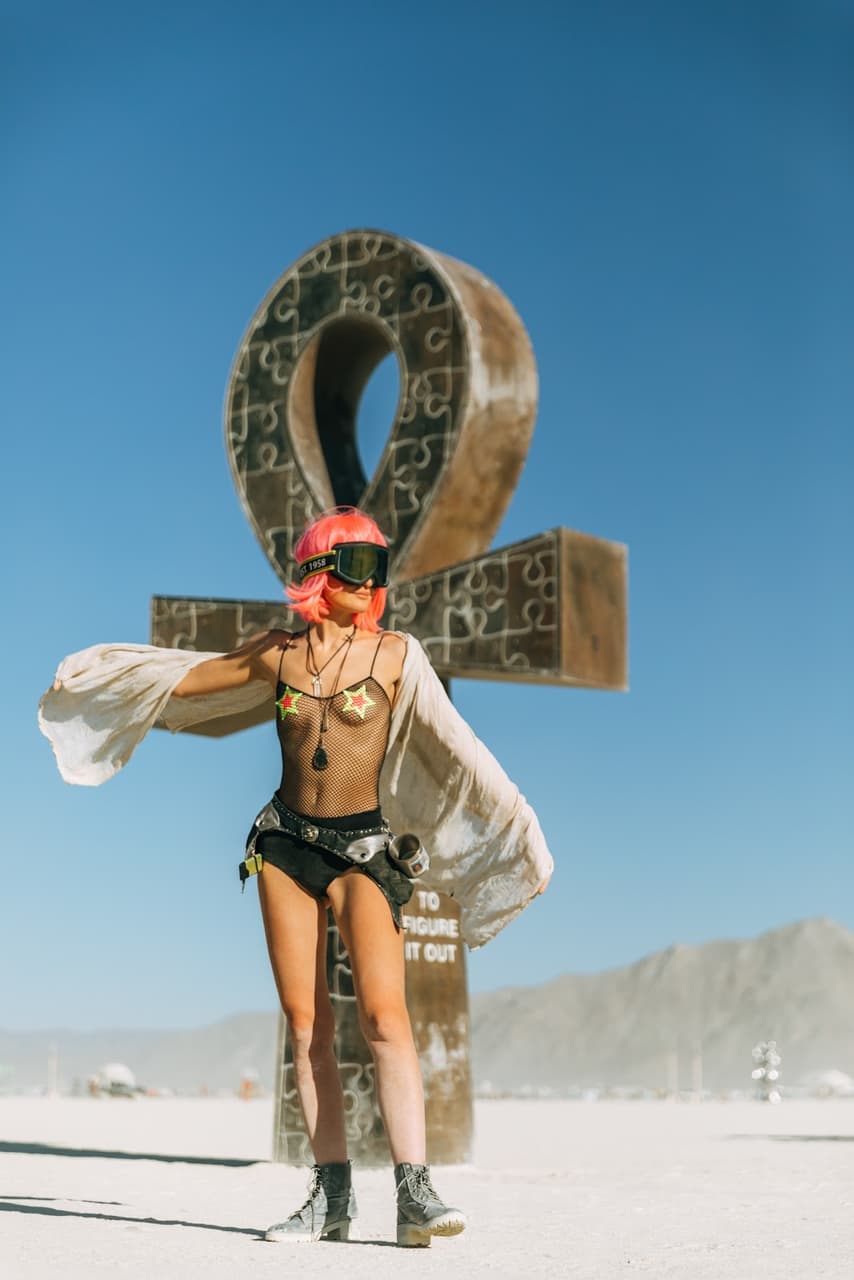 A woman with pink hair and goggles poses confidently in a revealing outfit in front of a massive ankh sculpture in a vast desert landscape.