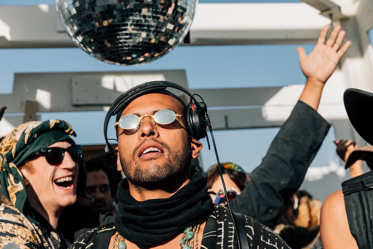 A man with headphones and sunglasses looks up at a disco ball, surrounded by other revelers in a bright outdoor setting.