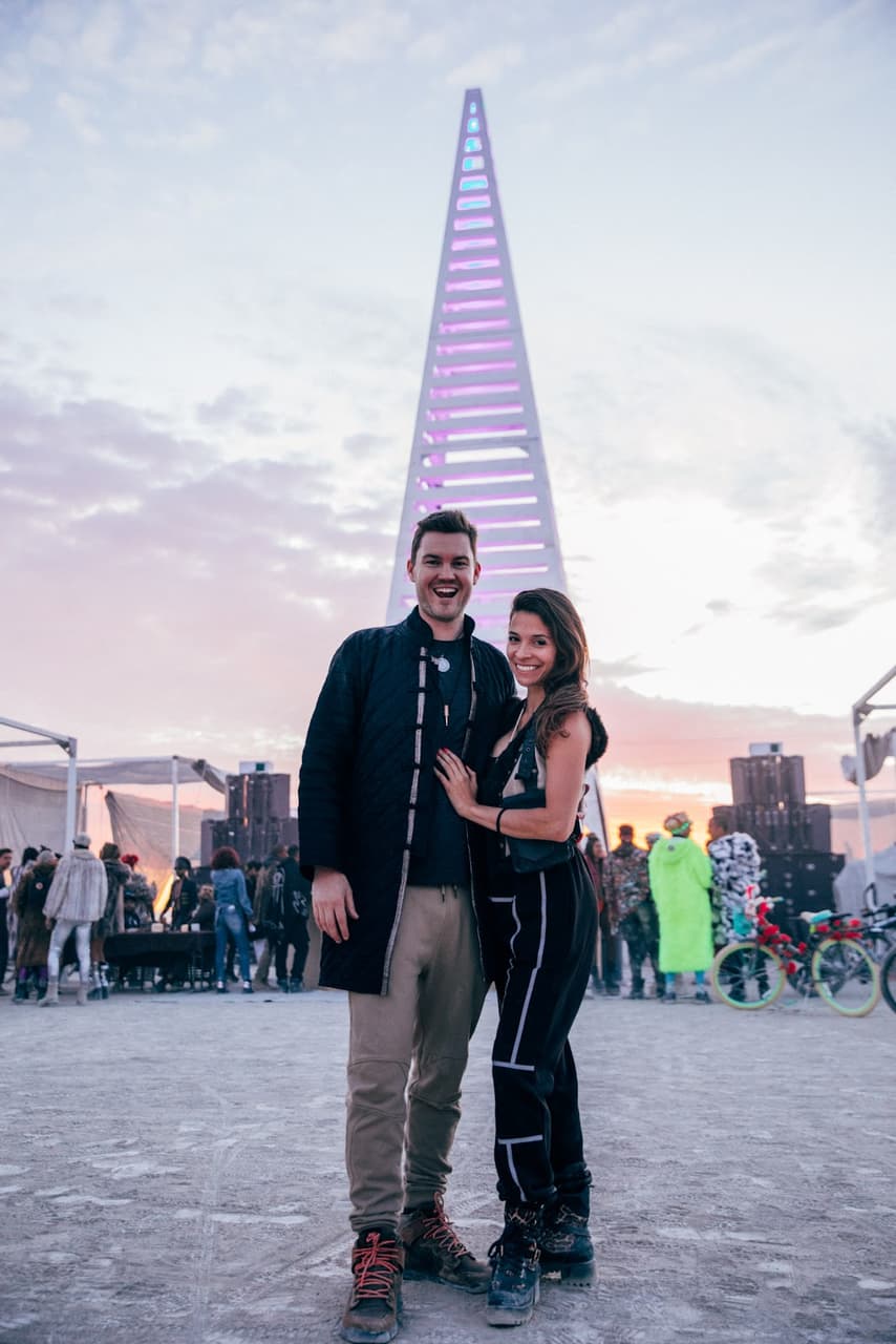 A smiling couple stands in front of a tall, glowing triangular art installation at an outdoor festival during sunset.