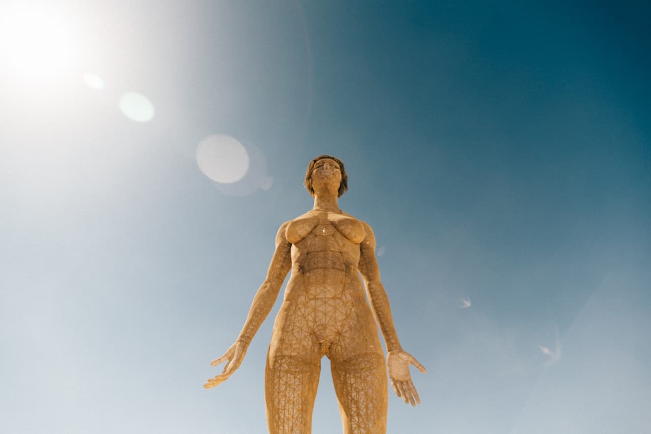 A towering, textured human-like sculpture stands against a bright blue sky with a prominent sun flare.
