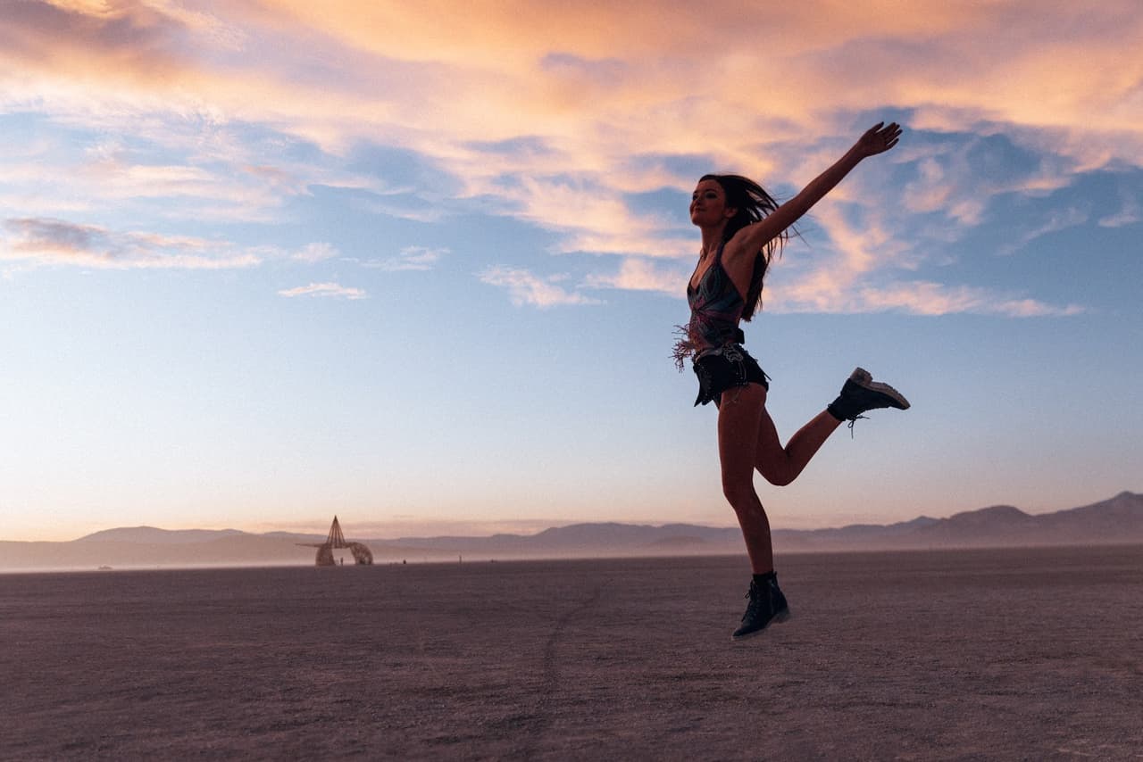 A woman joyfully leaps into the air against a vibrant sunset sky over a vast desert landscape.