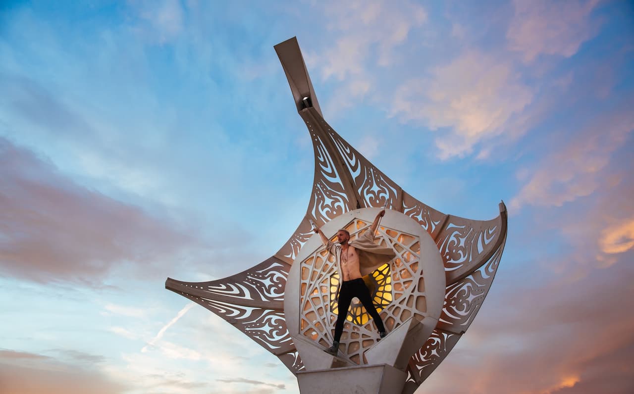 A person with arms outstretched stands dramatically within an ornate, manta-ray-like sculpture against a vibrant sunset sky.