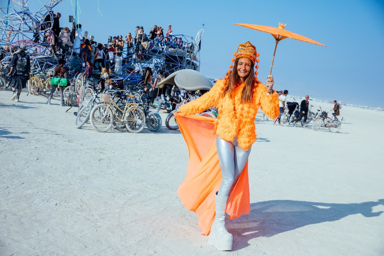 A woman in a vibrant orange outfit holds an orange parasol, walking across a dusty desert landscape with a large art installation and bicycl