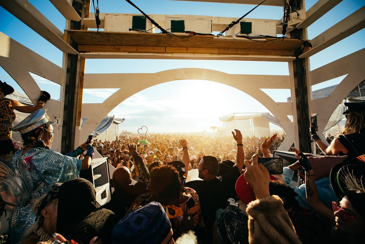 A vibrant crowd at an outdoor festival raises their hands towards a bright, backlighting sun framed by a white archway.