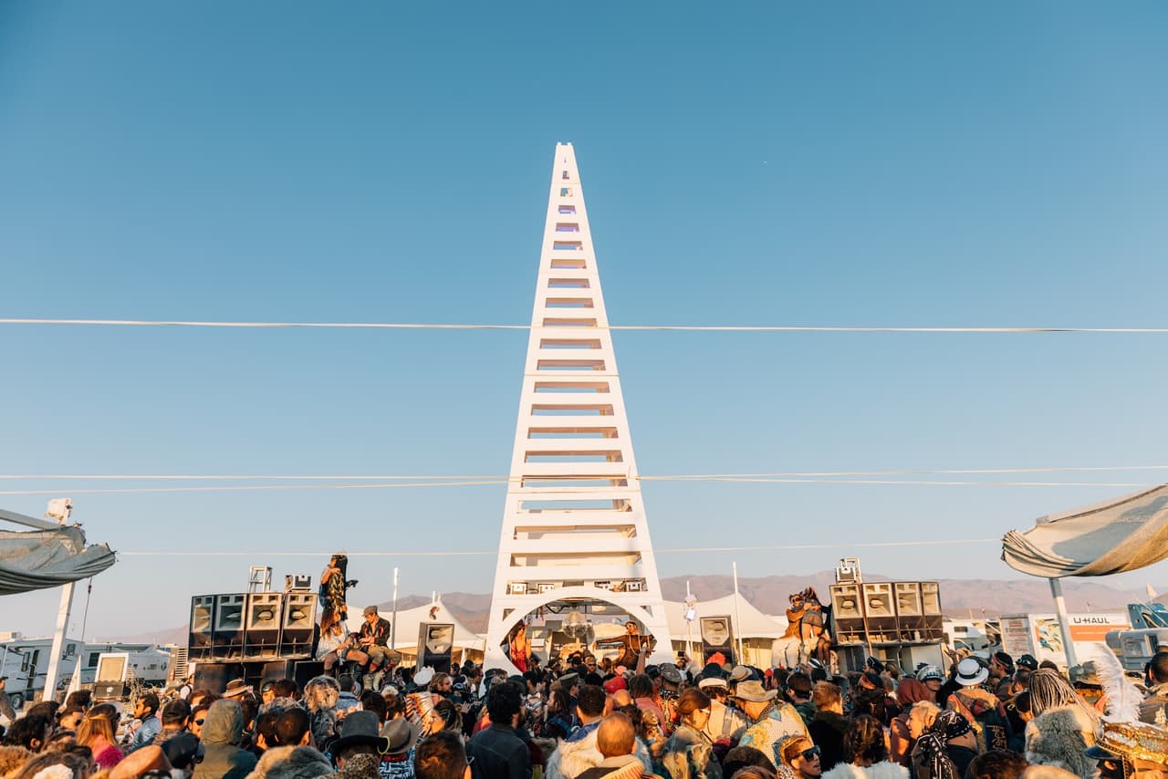 A large crowd gathers in front of a towering white triangular art installation at an outdoor festival under a clear blue sky.
