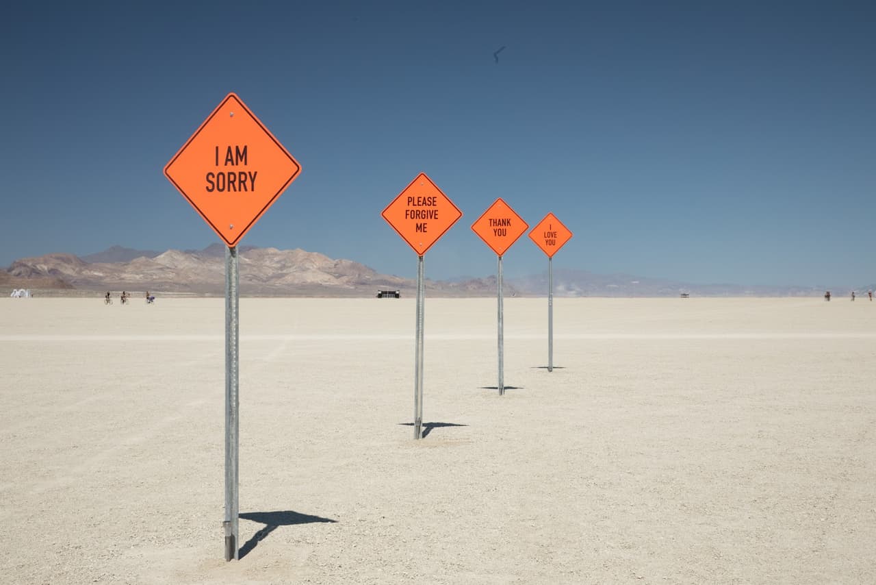 A line of orange diamond-shaped signs with messages like "I AM SORRY" and "PLEASE FORGIVE ME" stretches across a vast, flat desert landscape