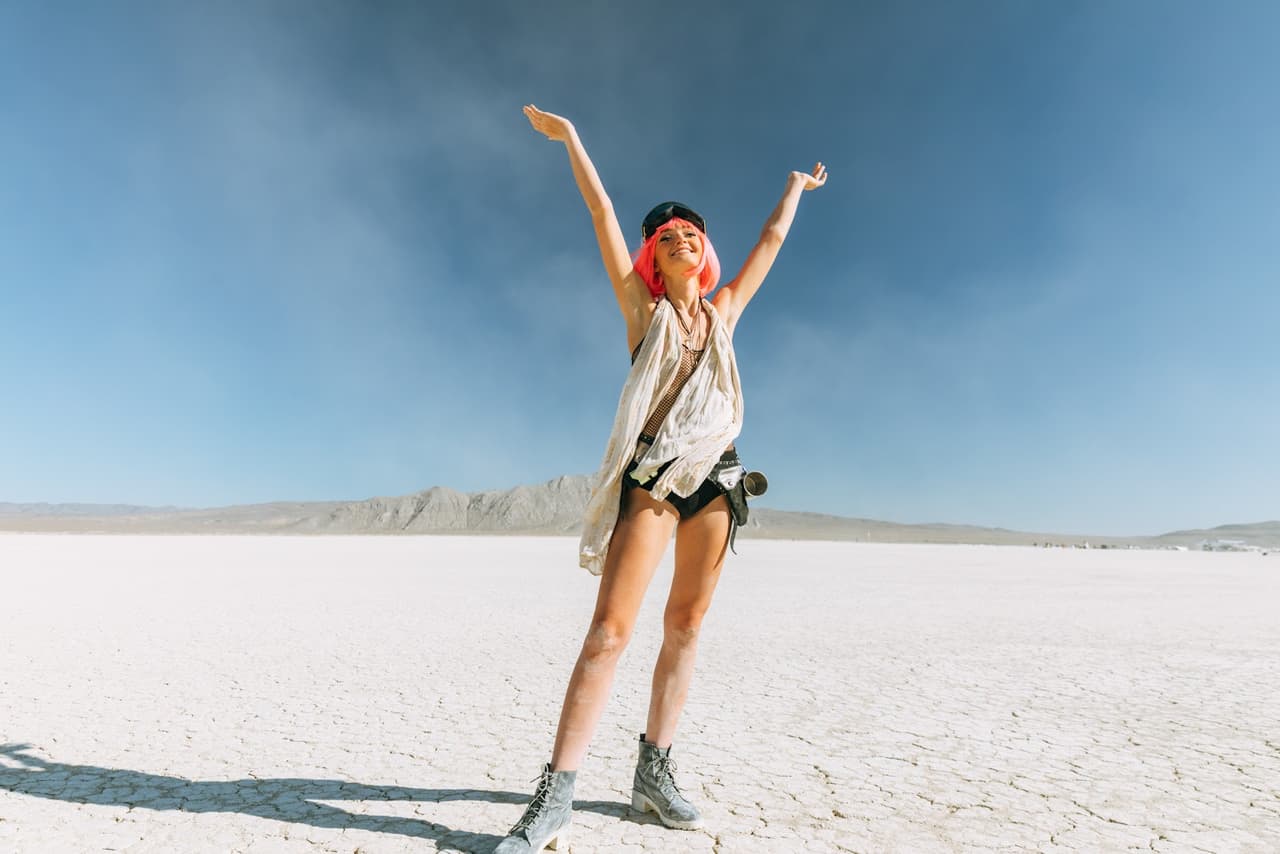 A joyful woman with vibrant pink hair stands with arms raised in a vast, cracked desert landscape under a clear blue sky.