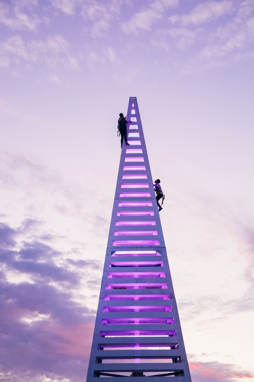Two people ascend a tall, glowing purple triangular structure against a vibrant purple and pink sky.