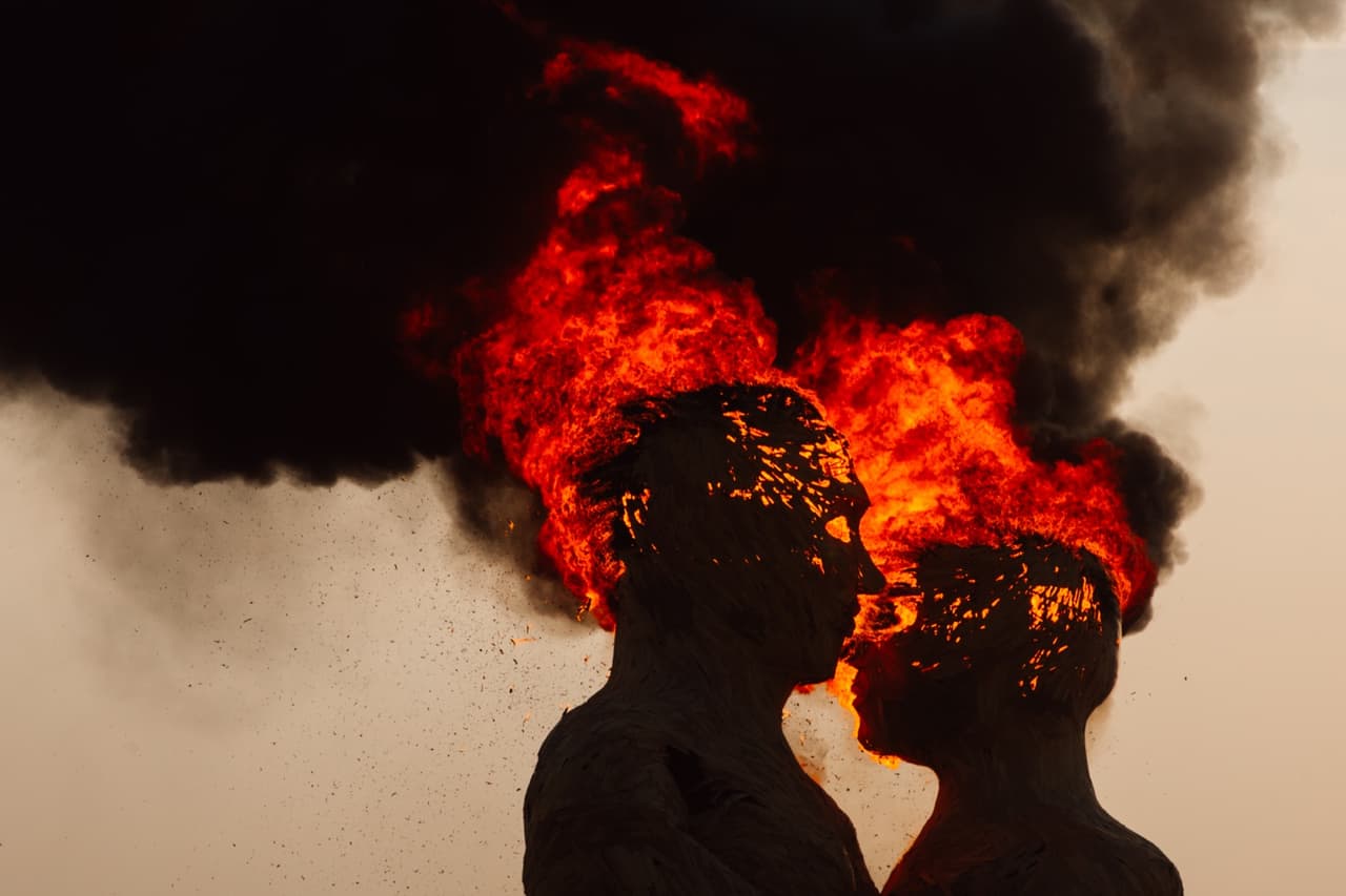 Two human-like figures, appearing to kiss, have their heads engulfed in dramatic flames and thick black smoke against a pale sky.