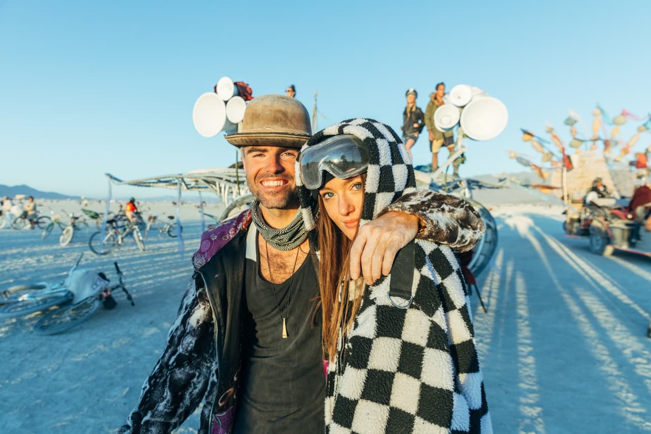 A smiling man in a hat and a woman in a black and white checkered hoodie pose together in a dusty desert landscape with art installations an