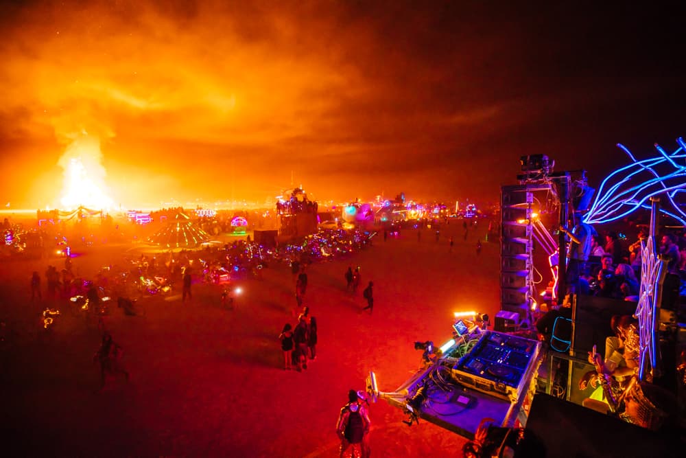 A vast outdoor night festival glows with an intense orange light from a massive distant fire under a dark, smoke-filled sky.