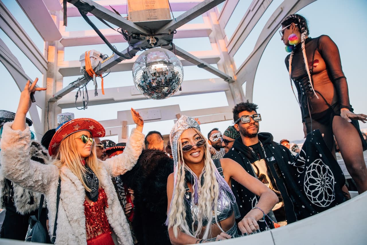 A group of festively dressed people dance and smile under a disco ball at an outdoor event.