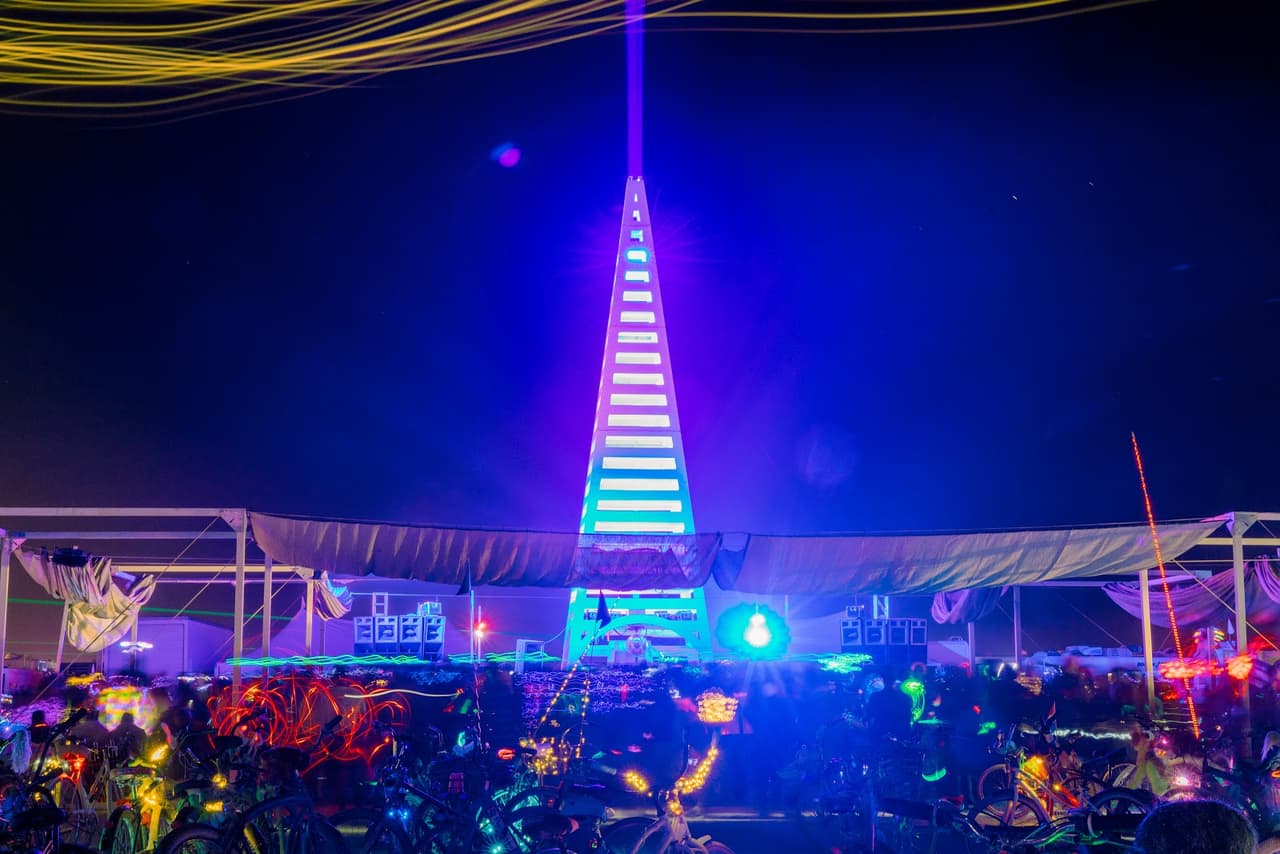 A towering, illuminated pyramid-like structure with a laser beam shooting into the night sky, surrounded by a vibrant crowd at an outdoor fe
