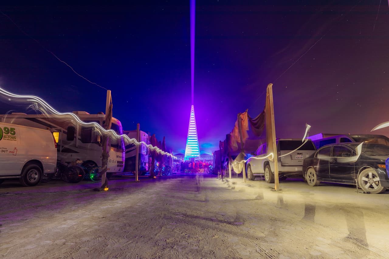 A vibrant purple and blue illuminated pyramid-like structure with a laser beam shooting skyward stands at the end of a dusty road lined with
