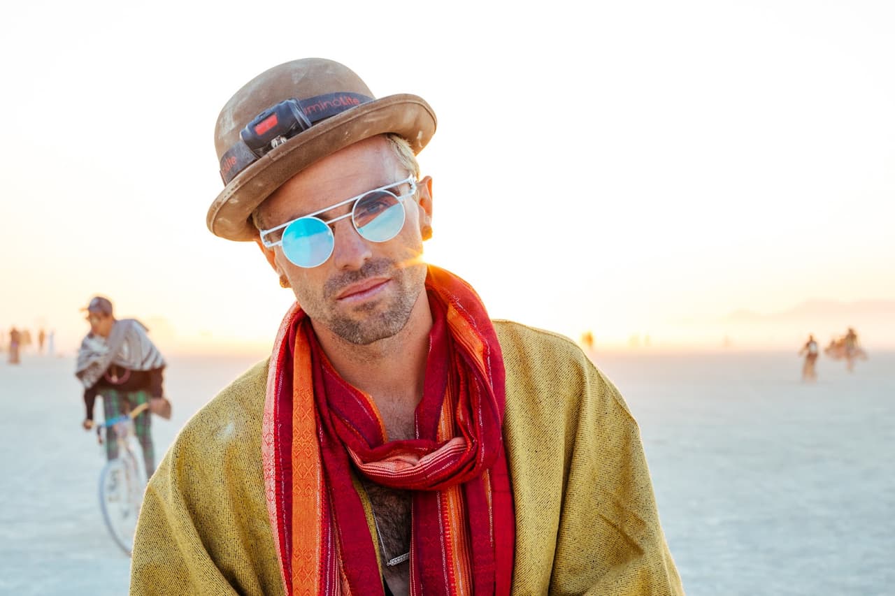 A man in a hat, reflective sunglasses, and a colorful scarf poses for a close-up portrait against a bright, hazy desert background at sunset