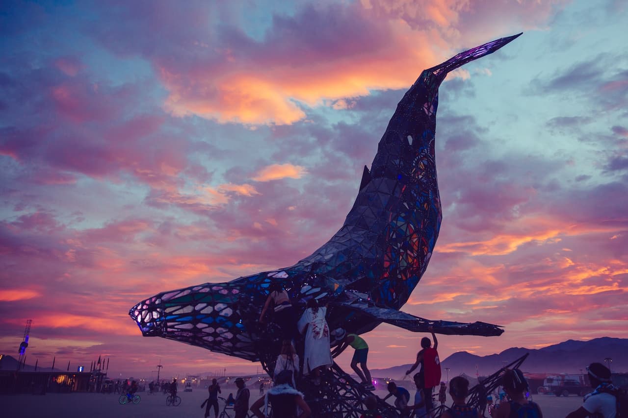 A massive, dark whale sculpture with illuminated patterns rises from a desert landscape against a vibrant sunset sky, with people climbing o