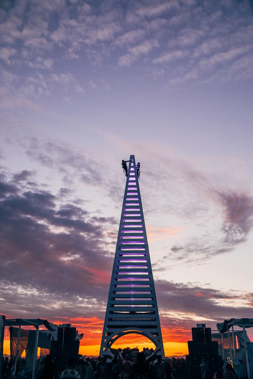 A tall, triangular art installation glows purple against a dramatic sunset sky with vibrant orange, red, and purple clouds.