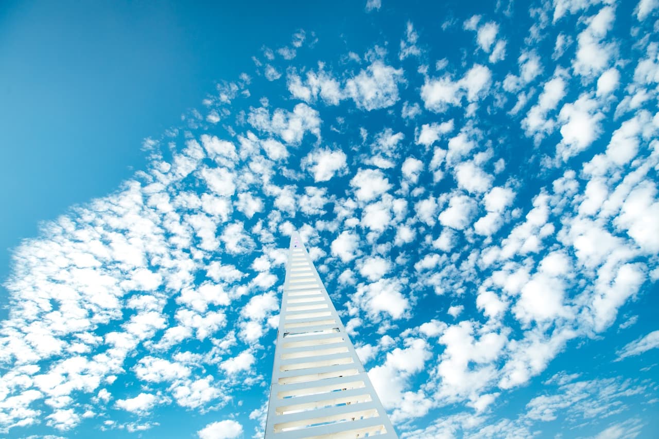 A tall, white ladder ascends vertically into a vibrant blue sky dotted with numerous small, fluffy white clouds.