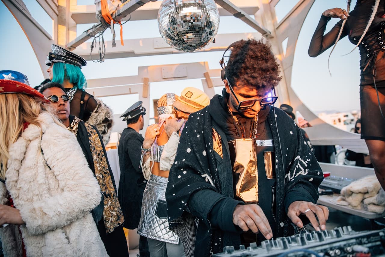 A DJ with a unique outfit and glowing headphones plays music at an outdoor festival, surrounded by a diverse crowd.