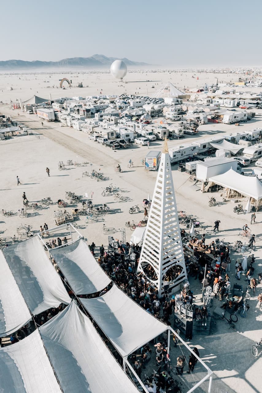 An aerial view captures a bustling desert festival with a distinctive white pyramidal tower surrounded by a large crowd and numerous tempora