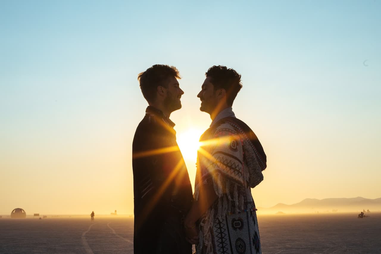 Two men are silhouetted against a brilliant sunset on a vast, flat landscape, holding hands and looking at each other.