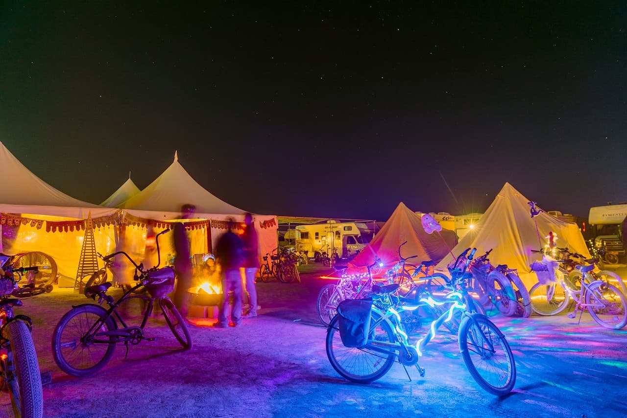 A vibrant night scene at a desert festival features illuminated bicycles, glowing tents, and people gathered under a starry sky.