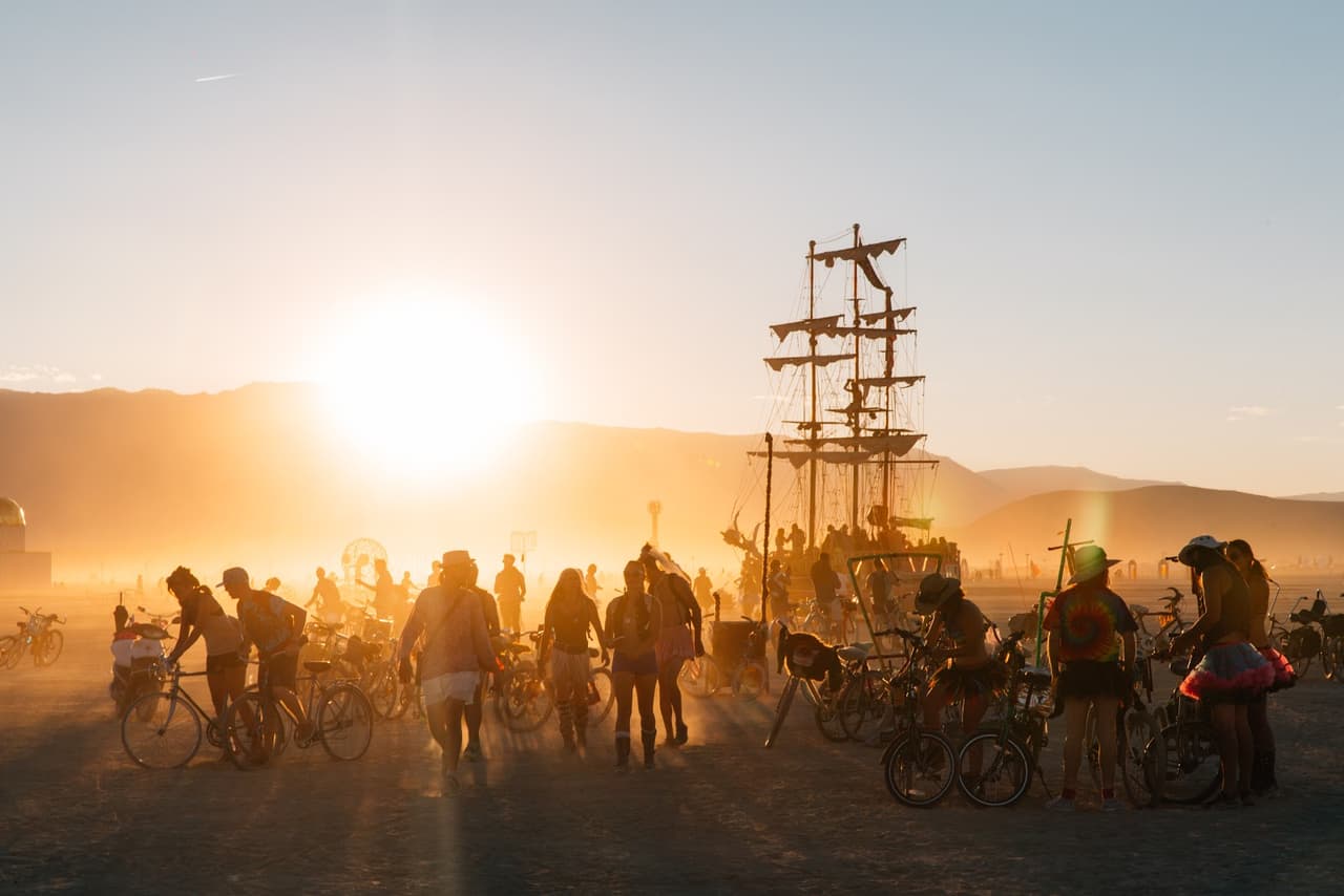 A large crowd of people and bicycles are silhouetted against a vibrant sunset in a dusty desert landscape, with a tall pirate ship art insta