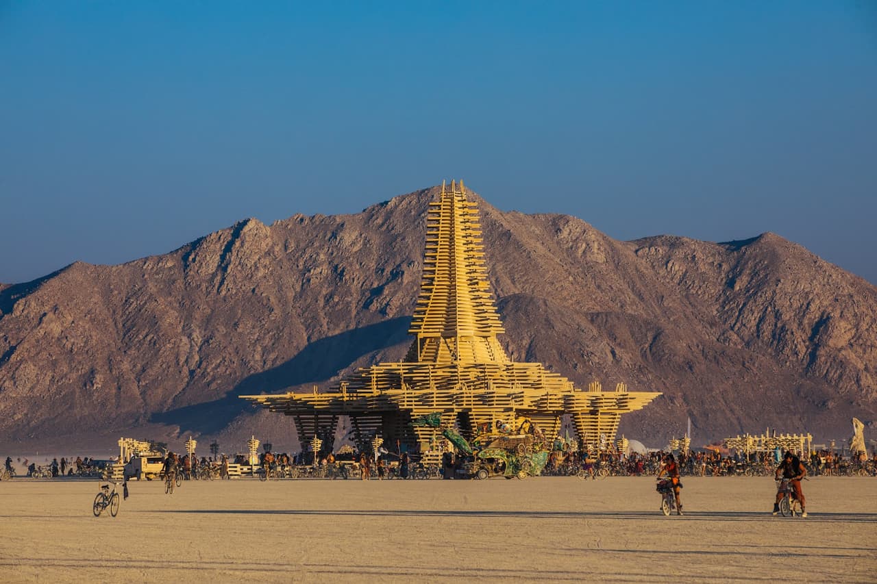 A towering, intricate wooden art installation stands prominently in a vast desert landscape with rugged mountains under a clear blue sky.