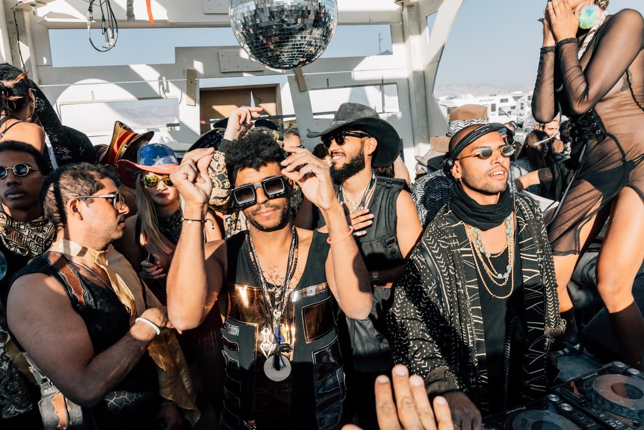 A diverse group of people in elaborate, festive attire dance under a disco ball at a vibrant outdoor event.