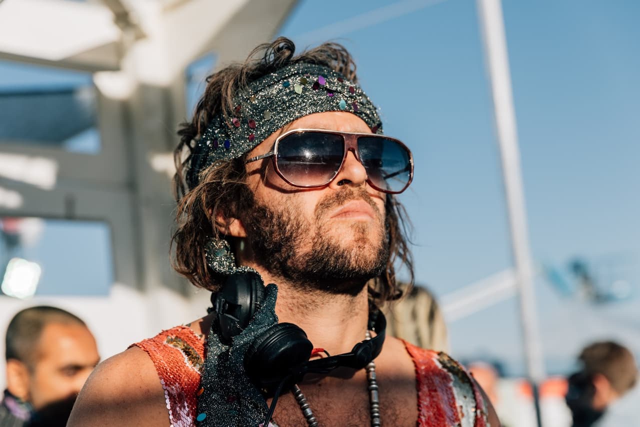 A man with a beard, sunglasses, and an elaborate headband looks upwards into the bright sunlight, with headphones around his neck.