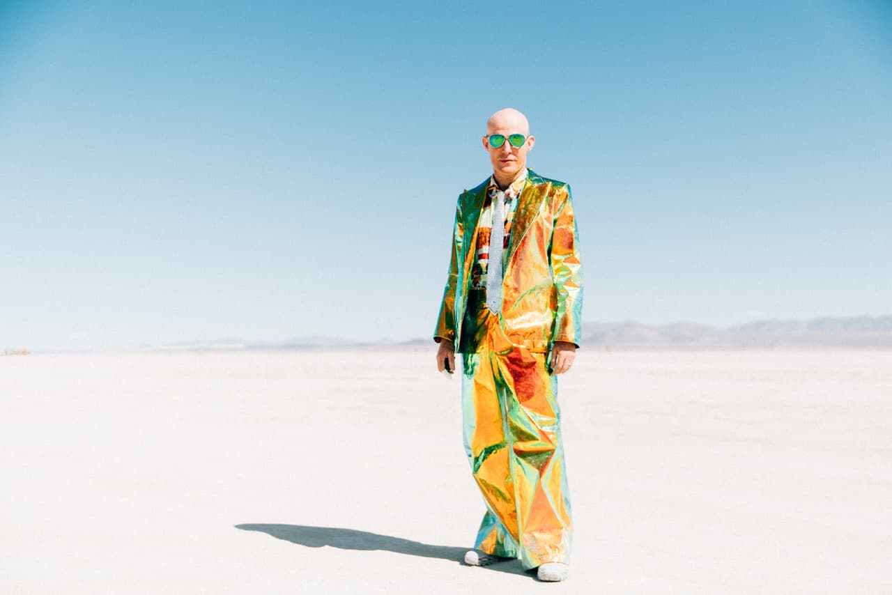 A bald person in a shimmering, iridescent geometric suit and green sunglasses stands on a vast, flat white desert landscape under a clear bl