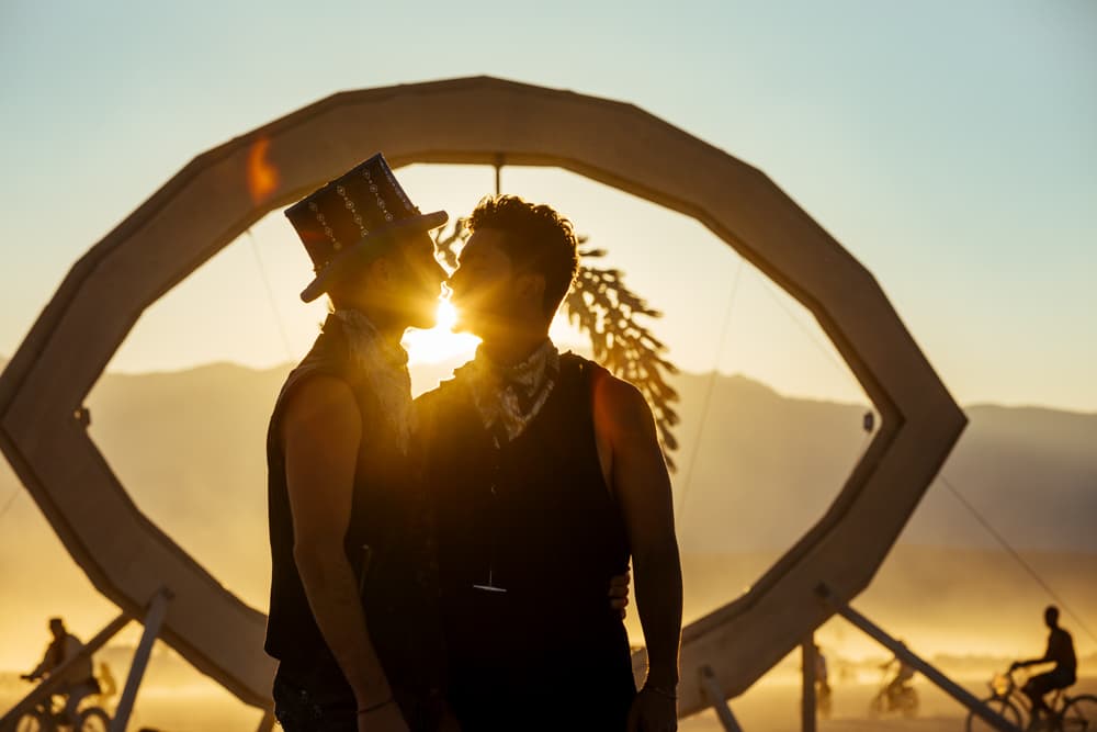 Two people in silhouette share a romantic kiss against a vibrant sunset, framed by a large art installation in a desert landscape.