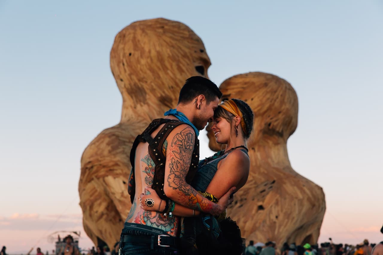 A tattooed couple embraces intimately in front of massive wooden sculptures under a golden hour sky.