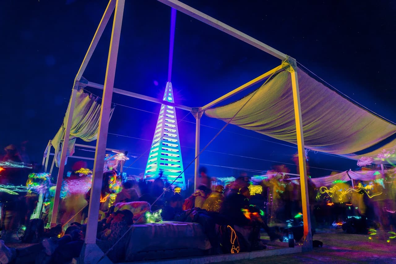 A vibrant nighttime scene shows a large, illuminated pyramid structure with a laser beam under a canopy filled with blurred figures.