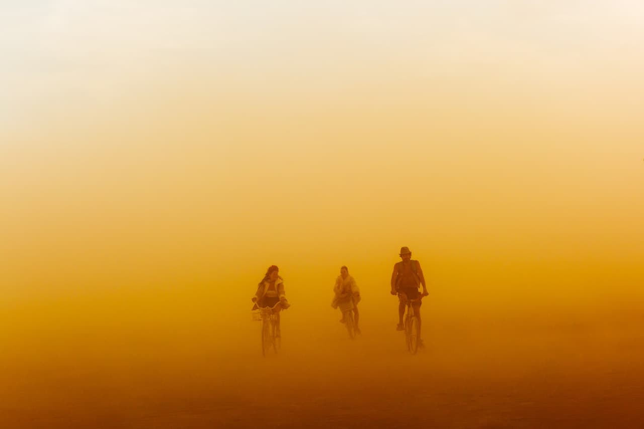 Silhouettes of four people cycle through a thick, orange-hued dust or fog under a gradient sky.