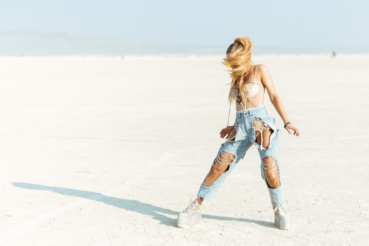 A woman with long blonde hair dramatically tossed to the side poses in heavily ripped jeans on a vast, bright white salt flat.