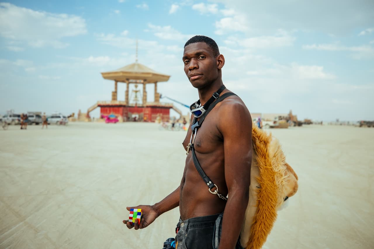 A shirtless man with a Rubik's cube stands in a vast desert landscape, wearing goggles around his neck and a furry backpack.