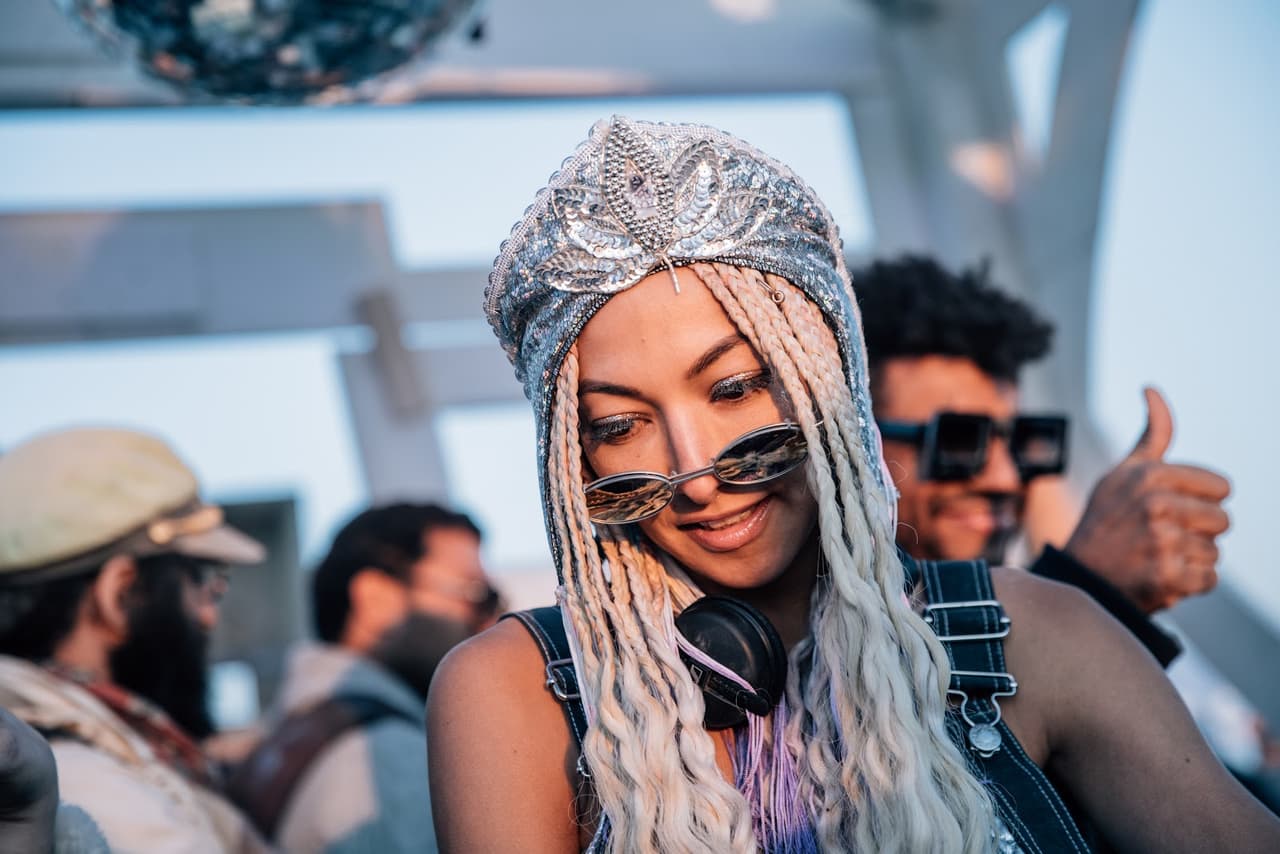 A woman with blonde dreadlocks and a sparkling silver turban smiles while wearing sunglasses and headphones at an outdoor event.