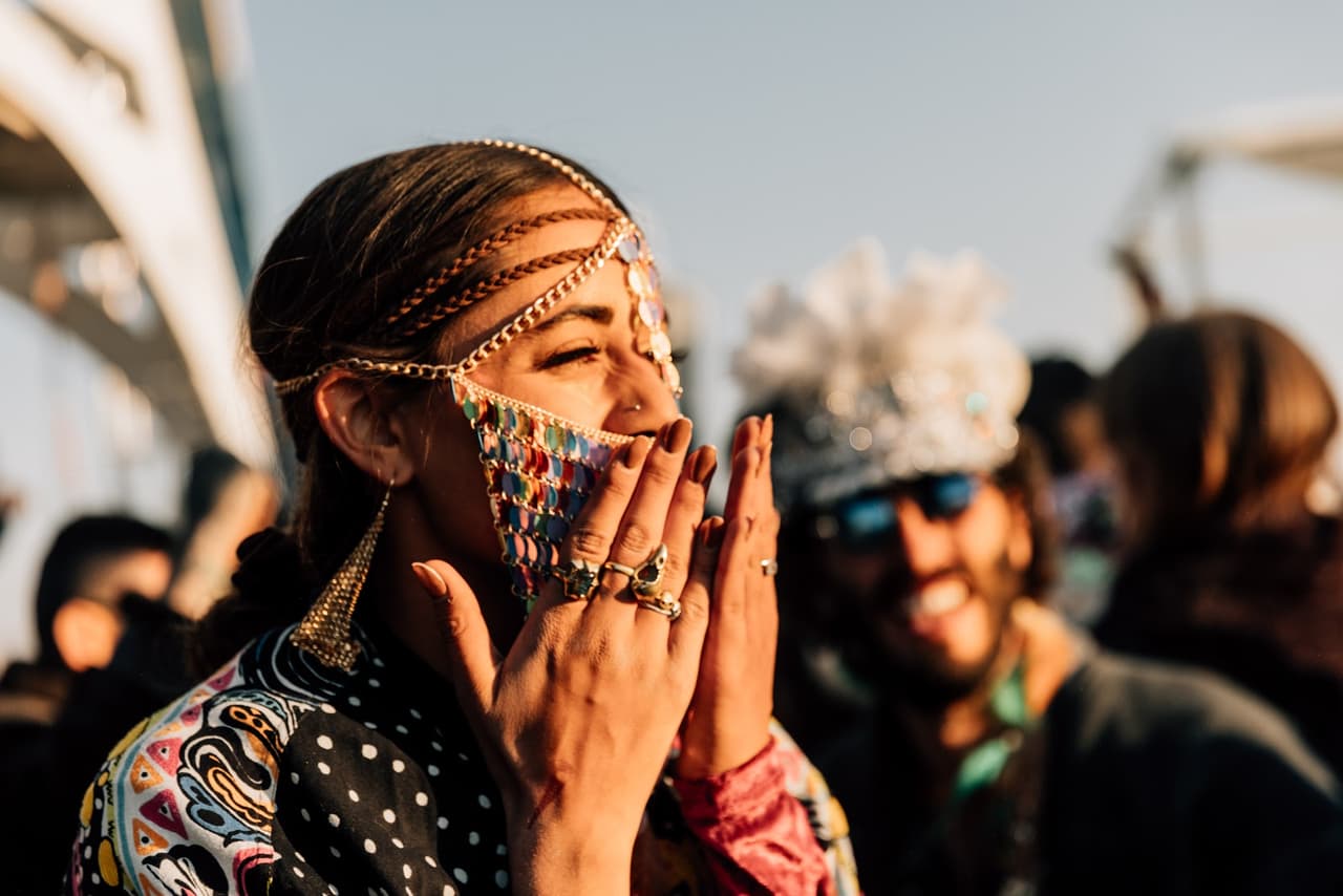 A woman with an elaborate headpiece and colorful beaded face covering laughs joyfully, illuminated by golden hour sunlight.