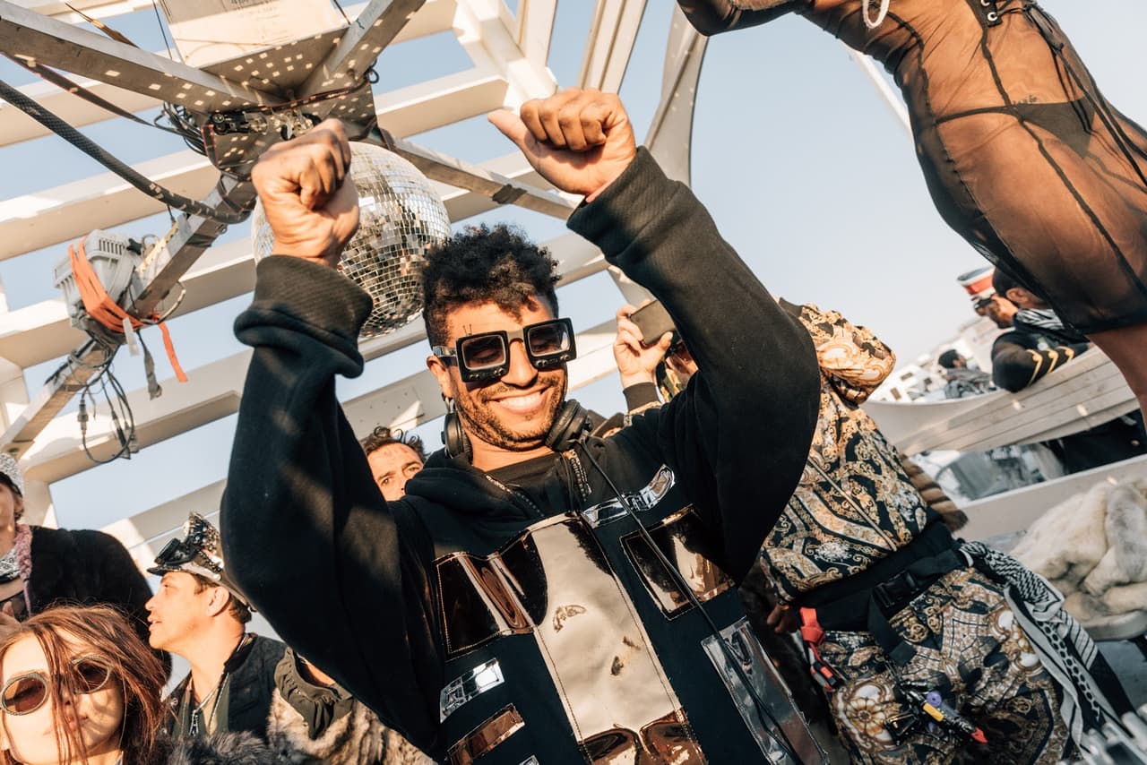 A man in sunglasses and a black metallic hoodie joyfully dances with his arms raised at an outdoor event.