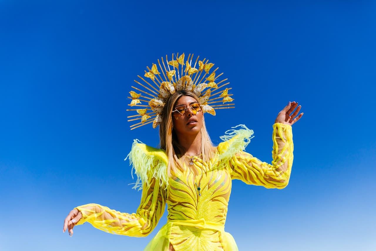 A woman in a vibrant yellow outfit and an elaborate golden butterfly headdress poses against a brilliant blue sky.