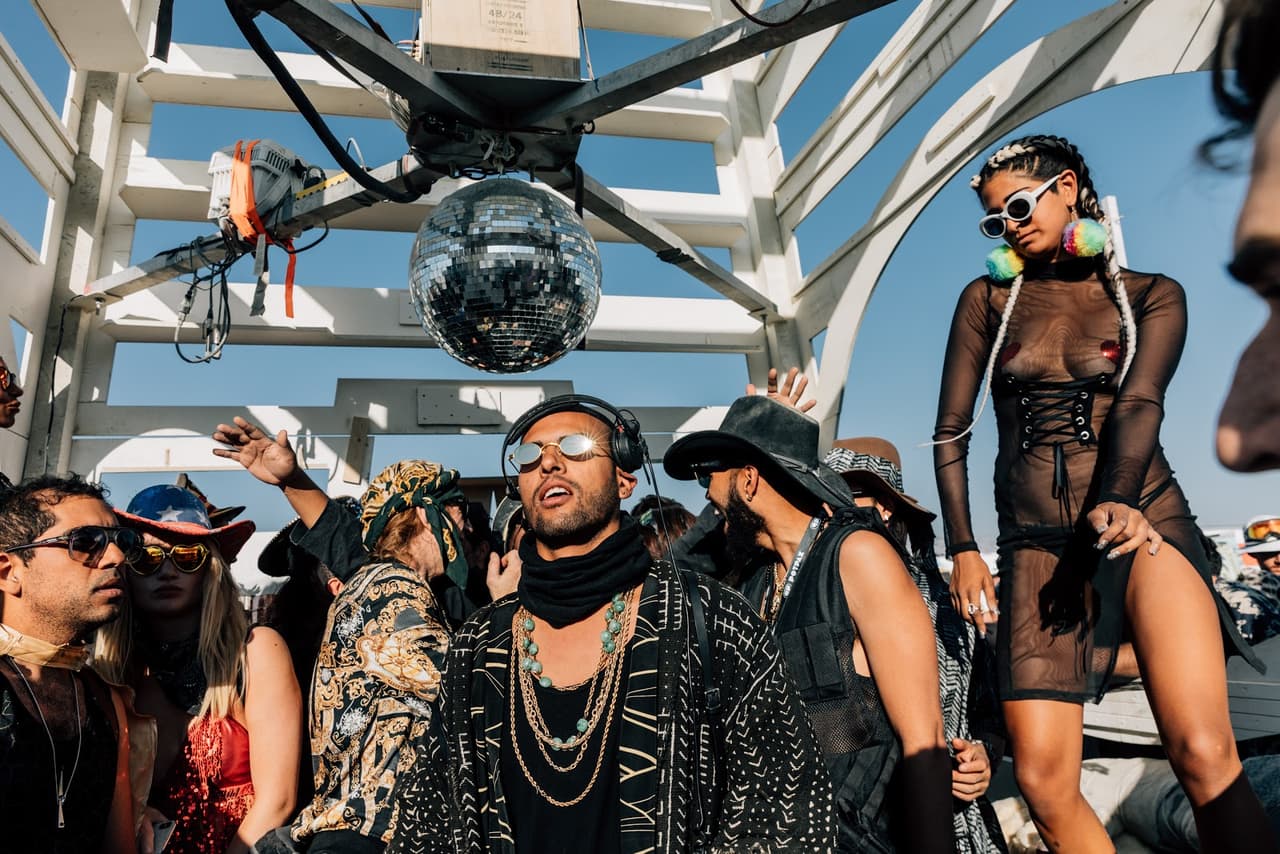 A man with sunglasses and headphones sings into a microphone amidst a vibrant outdoor festival crowd under a clear blue sky, with a disco ba