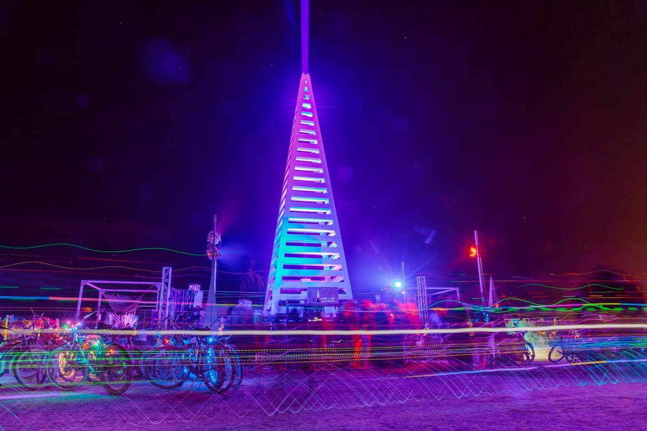 A vibrant, multi-colored light-up pyramid art installation stands tall at night, surrounded by streaks of light from moving objects.