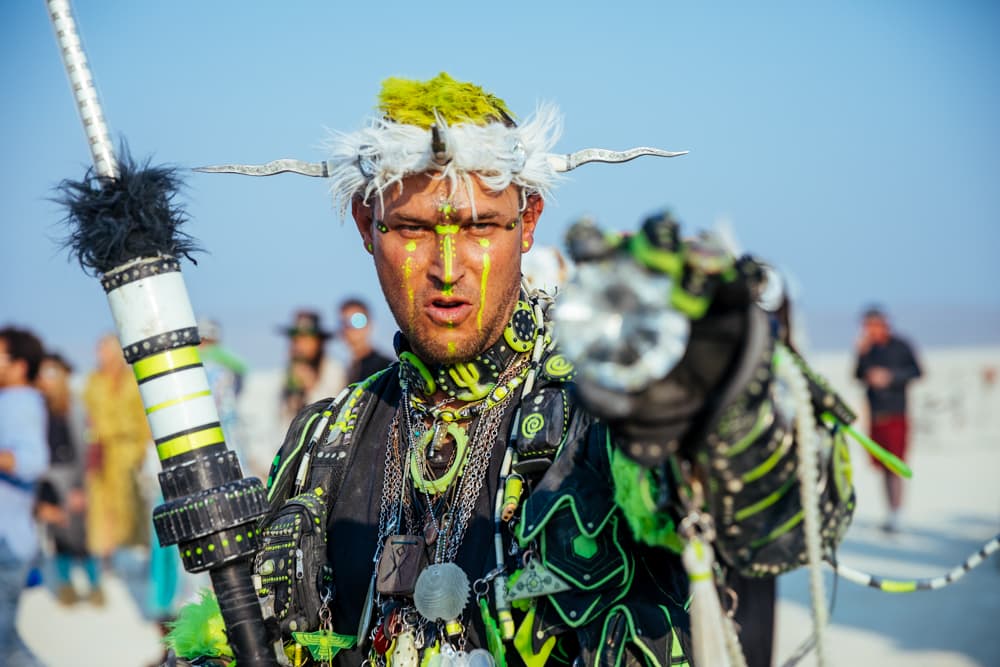 A person in an elaborate neon green and black costume with face paint and a horned headdress points a glowing object directly at the viewer