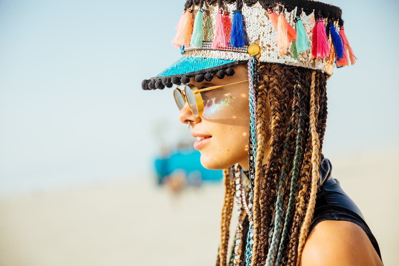 A woman with elaborate braids, a sequined hat with colorful tassels, and glitter on her face smiles in profile under bright sunlight.